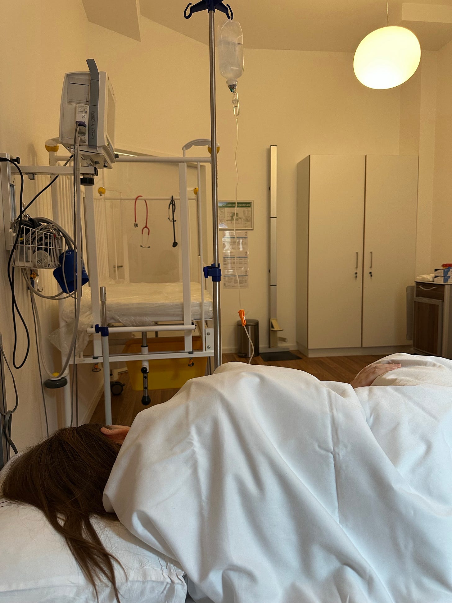 A patient lying on a hospital bed covered with a white blanket, connected to an IV stand, with medical equipment and hospital furniture visible in the background.