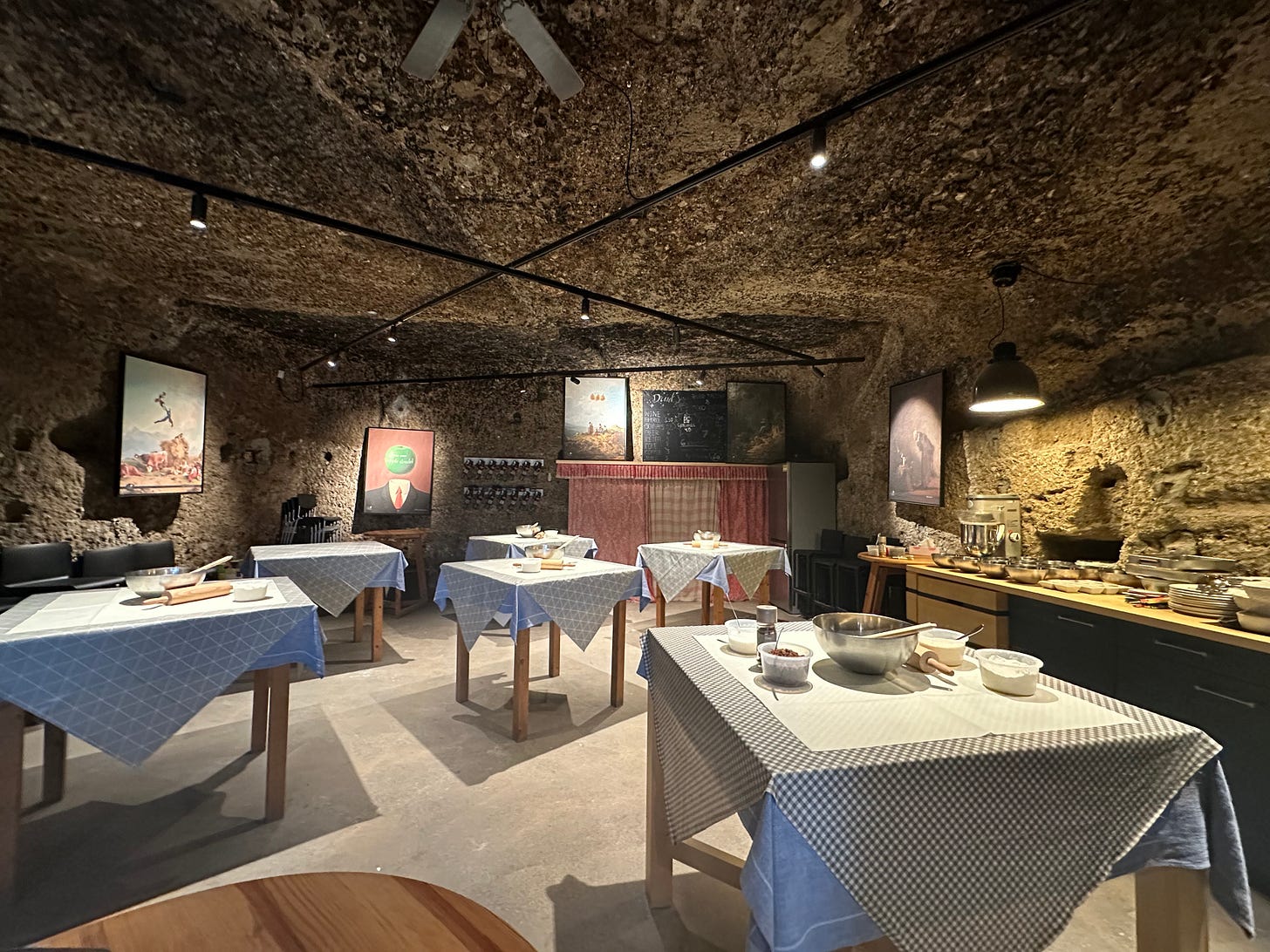 Interior of a cave restaurant with rustic decor, featuring several tables with blue tablecloths, culinary preparation items, and artwork on the stone walls.