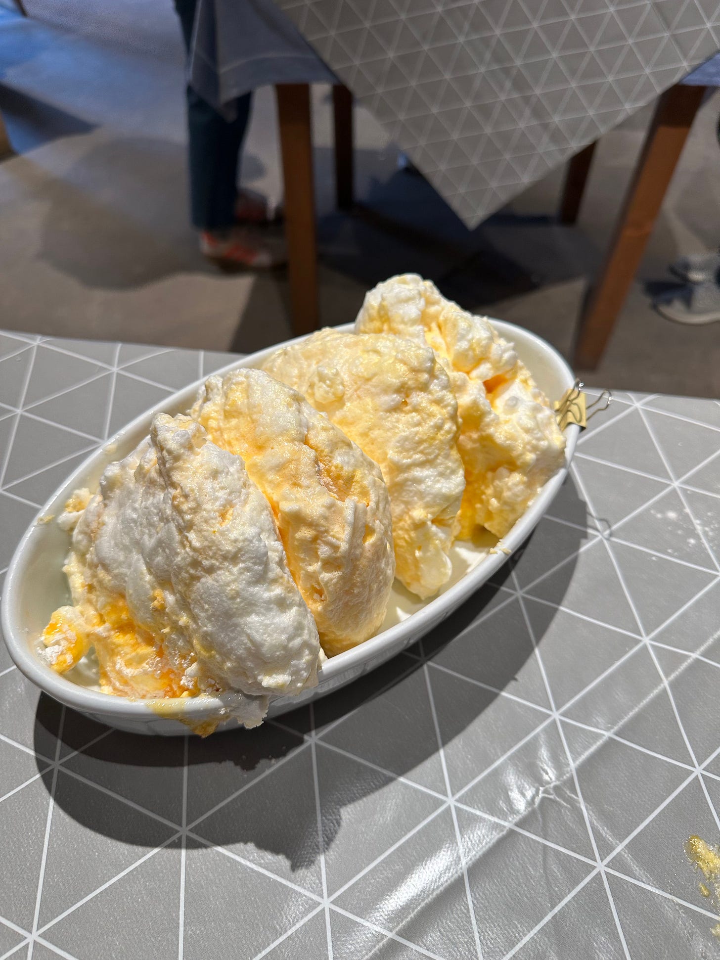A white dish filled with fluffy, yellow-tinted dessert resembling meringue or whipped cream, placed on a geometric-patterned table.