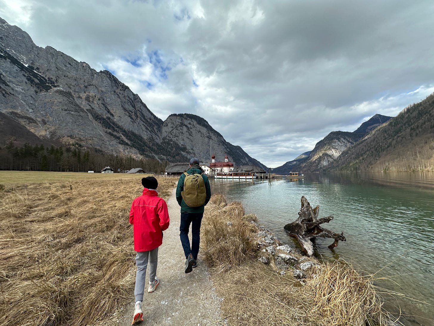A child in a red jacket and an adult walking along a riverside path, surrounded by mountains and cloudy skies.