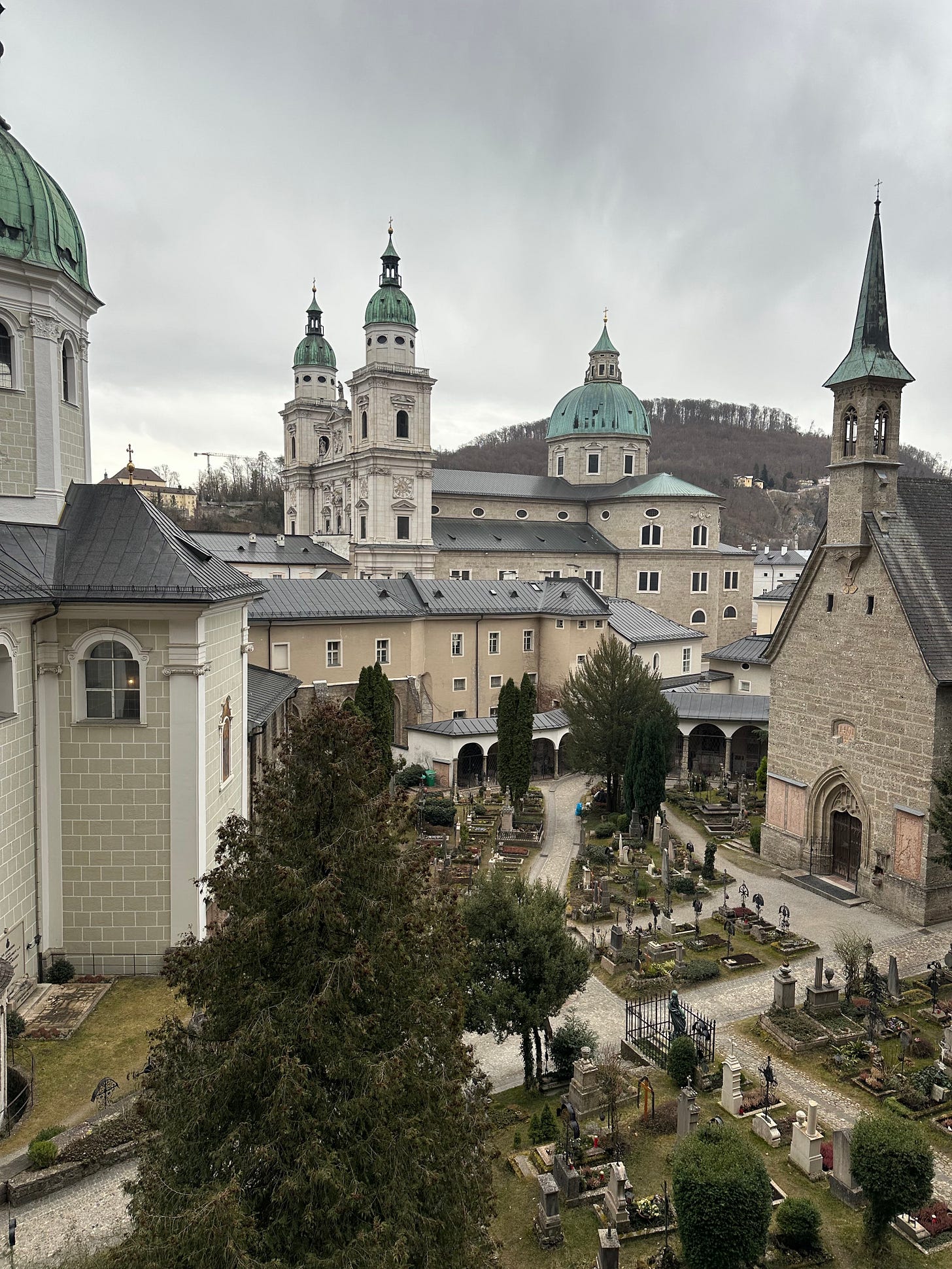 View of a historical church complex with domed roofs and bell towers, surrounded by a cemetery featuring numerous graves and green trees under a cloudy sky.