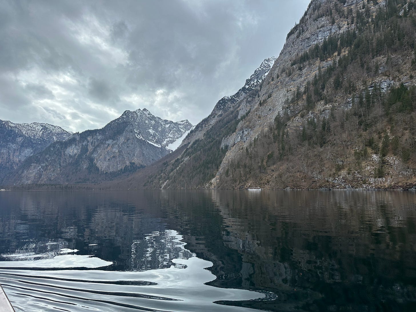 A serene mountain lake surrounded by steep, rocky mountains under a cloudy sky, with a small boat visible on the water and reflections of the landscape in the calm surface.