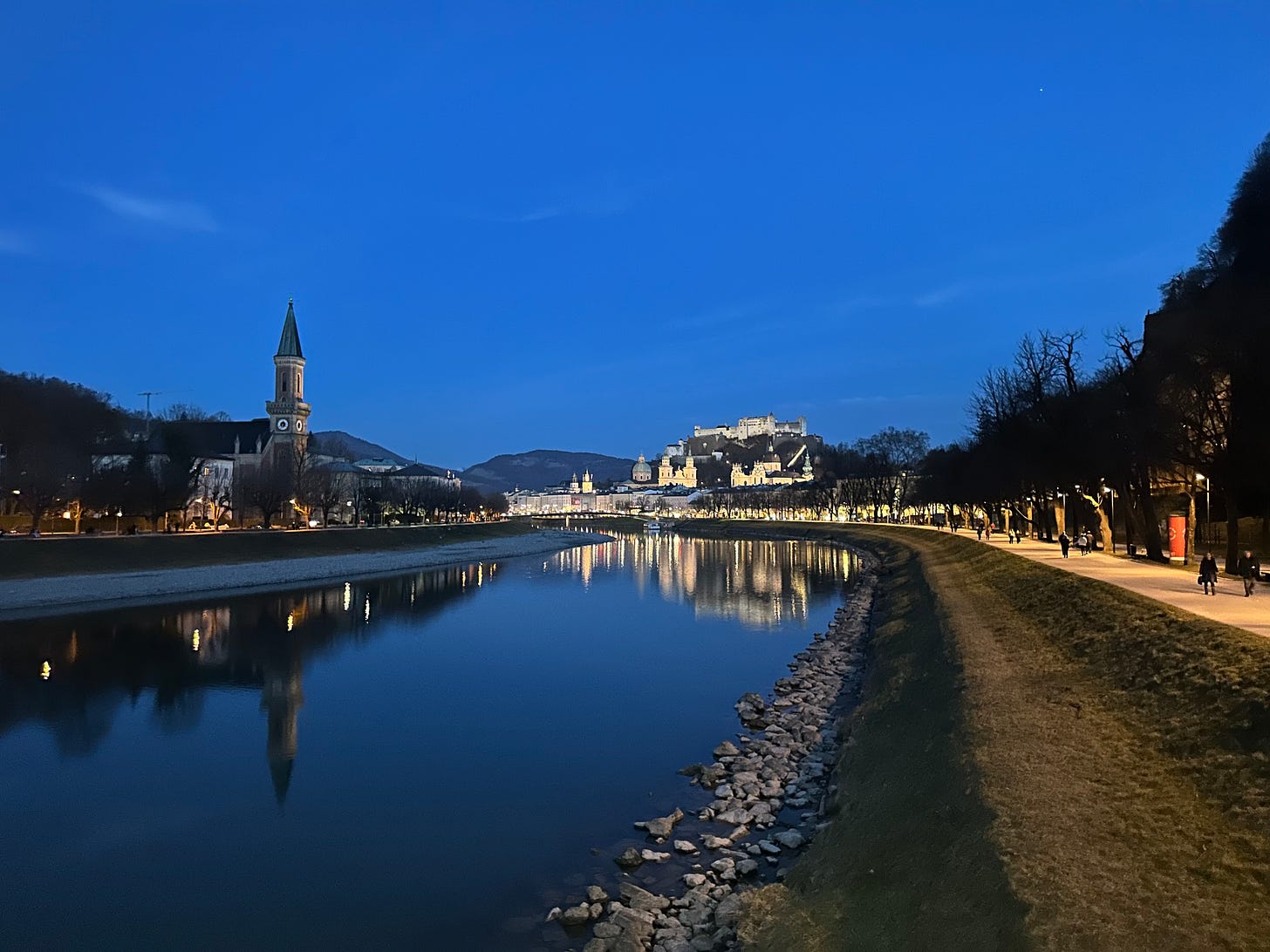 A dusk view of a riverbank with a church tower and illuminated buildings in the background, reflecting in the calm water.
