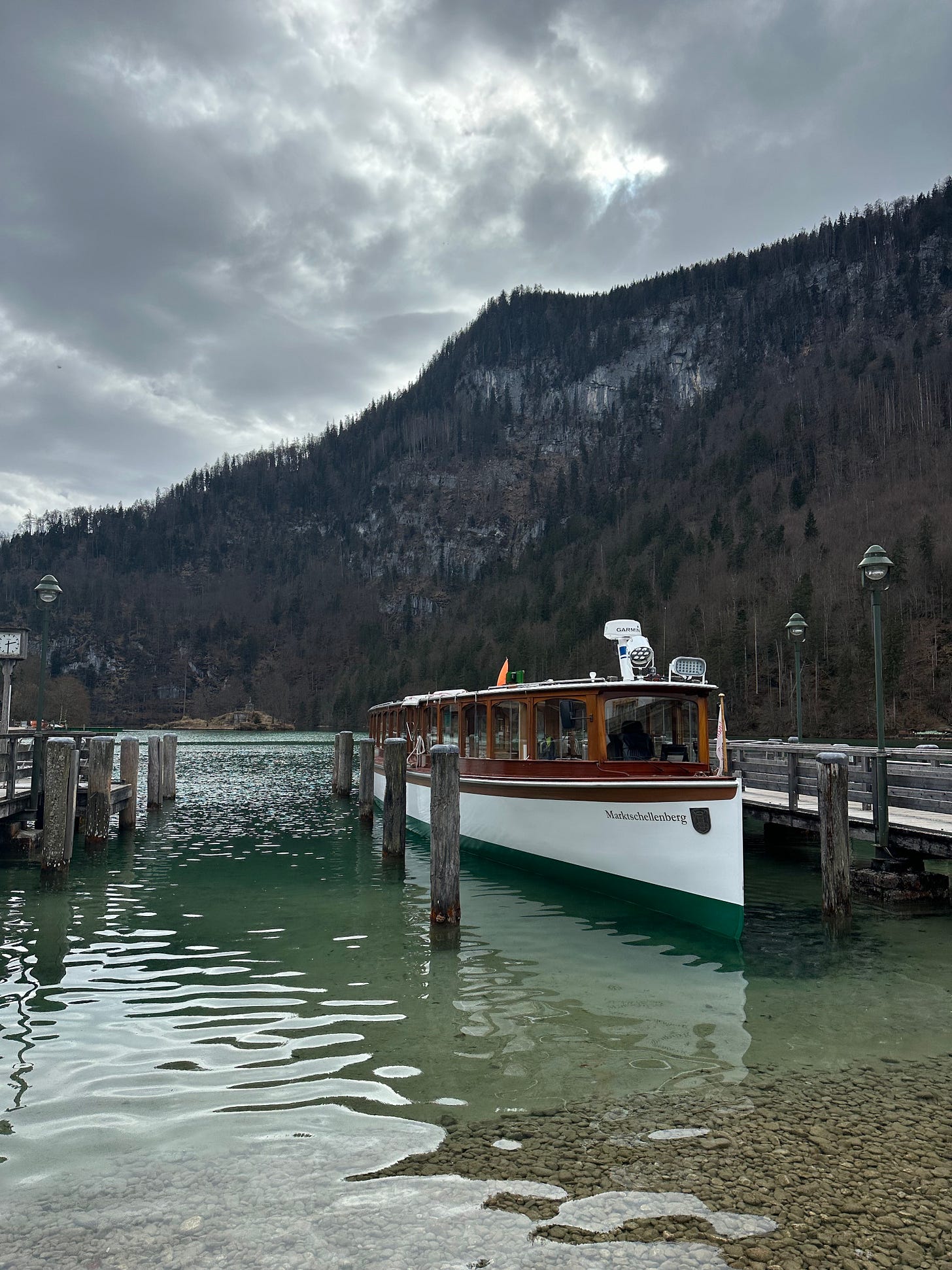 A wooden boat docked at a pier on a lake surrounded by mountains, under a cloudy sky.